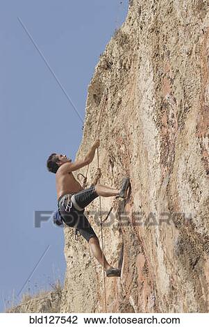 Stock Image - Male rock climber scaling rock face secured by ropes low angle view. Fotosearch