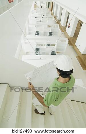 Stock Image - Man wearing hard hat standing in new office holding plans. Fotosearch