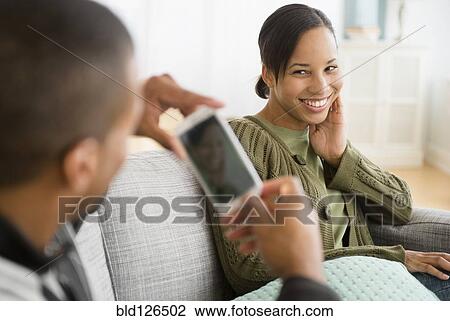 Stock Image - Man with camera phone photographing girlfriend on sofa. Fotosearch
