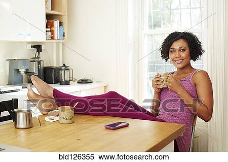 Stock Photography - Mixed race woman drinking coffee with feet up in kitchen. Fotosearch