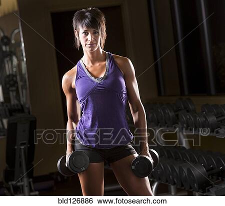 Stock Photograph - Portrait of confident Caucasian woman holding dumbbells in gym. Fotosearch