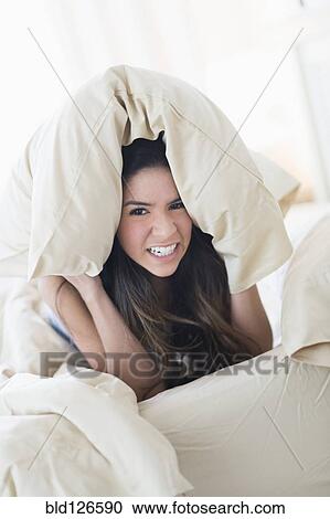 Stock Image - Portrait of frustrated Hispanic woman under pillow in bed. Fotosearch