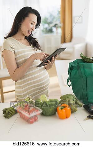 Stock Photograph - Pregnant Japanese woman using digital tablet in kitchen. Fotosearch