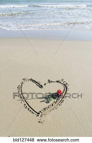 Stock Photo - Rose laid on heart drawn in sand. Fotosearch