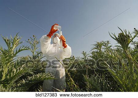 Stock Image - Woman in boiler suit standing in field pouring liquid into test tube. Fotosearch