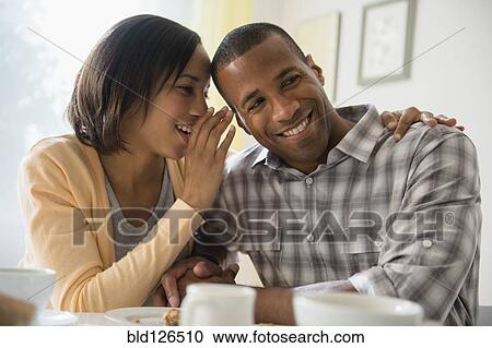 Stock Image - Woman whispering in boyfriend's ear at table. Fotosearch