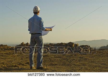 Stock Image - Young engineer with plan on construction site rear view. Fotosearch