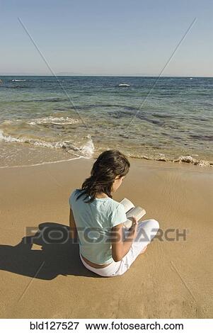 Stock Photo - Young woman sitting on beach reading book rear view. Fotosearch