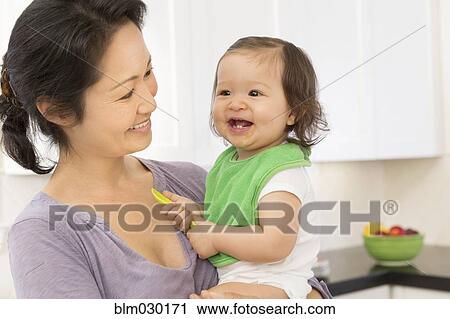 Stock Image - Mother holding baby girl in kitchen. Fotosearch