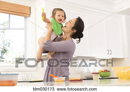Stock Image - Mother holding baby girl in kitchen. Fotosearch