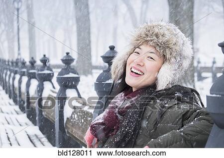 Stock Image - Asian woman sitting on park bench in snow. Fotosearch