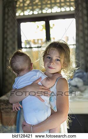 Stock Image - Caucasian girl carrying baby boy in bedroom. Fotosearch