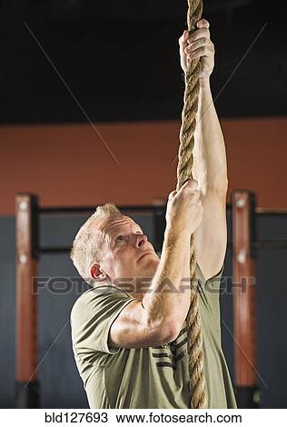 Stock Image - Caucasian man climbing rope in gym. Fotosearch