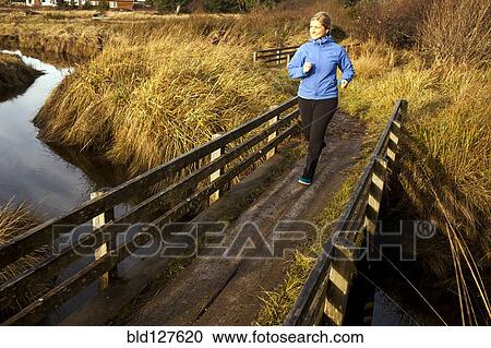 Stock Image - Caucasian woman jogging on wood bridge over creek. Fotosearch