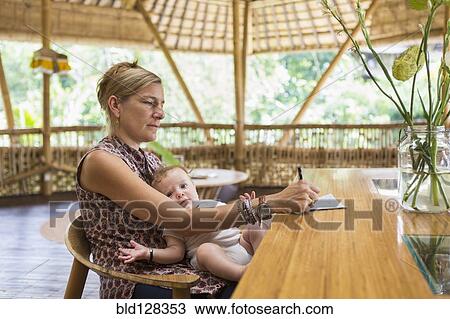 Stock Image - Caucasian woman with baby writing at table. Fotosearch