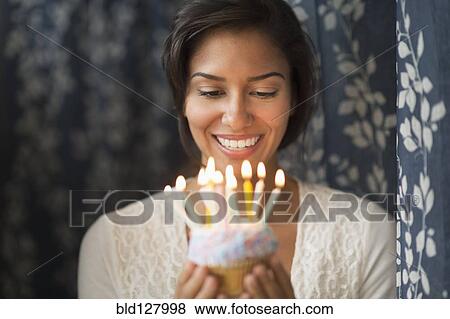 Stock Photo - Hispanic woman looking down at lit candles on birthday cupcake. Fotosearch
