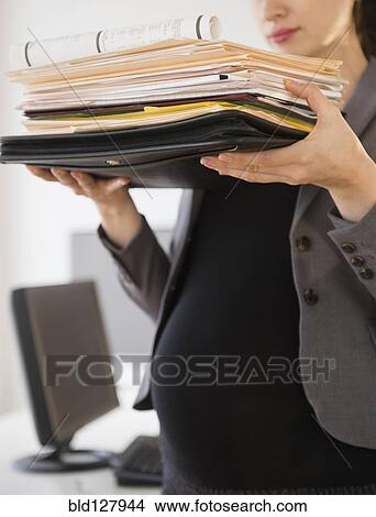 Picture - Pregnant Caucasian businesswoman carrying folders in office. Fotosearch