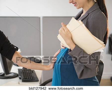 Stock Image - Pregnant Caucasian businesswoman showing off belly in office. Fotosearch