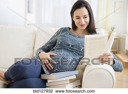 Stock Image - Pregnant Caucasian woman reading baby books on sofa. Fotosearch