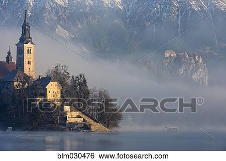 Stock Photograph - Fog shrouding island and mountains on Lake Bled at sunrise, Bled, Gorenjska, Slovenia. Fotosearch