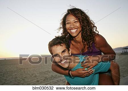 Stock Image - Man carrying girlfriend piggyback on beach. Fotosearch