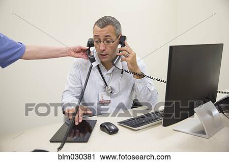 Stock Image - Mixed race doctor answering two phones and using digital tablet in office. Fotosearch