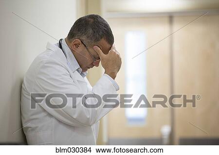 Picture - Mixed race doctor rubbing his forehead in hospital corridor. Fotosearch