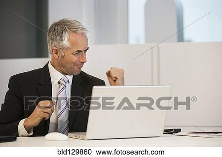 Stock Image - Businessman cheering at desk in office. Fotosearch