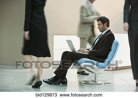 Businessman working on laptop in busy lobby area View Large Photo Image Stock Image - Businessman working on laptop in busy lobby area. Fotosearch