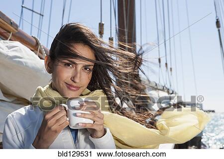 Stock Image - Caucasian woman drinking coffee on sailboat. Fotosearch