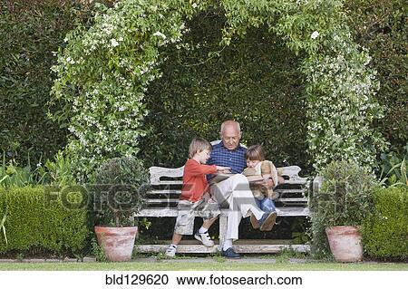 Stock Image - Grandfather reading book to grandchildren on garden bench. Fotosearch