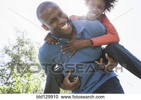Stock Image - Man carrying girlfriend piggyback in park. Fotosearch