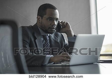 Picture - Mixed race businessman talking on cell phone at desk. Fotosearch