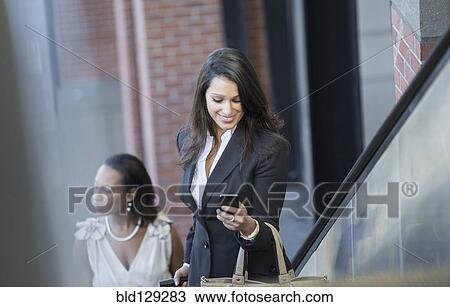 Stock Image - Mixed race businesswoman using cell phone on escalator. Fotosearch