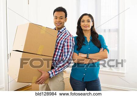 Picture - Mixed race couple carrying cardboard boxes in new home. Fotosearch