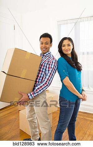 Stock Image - Mixed race couple carrying cardboard boxes in new home. Fotosearch