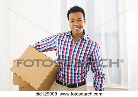 Stock Photo - Mixed race man carrying cardboard box in new home. Fotosearch
