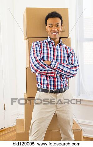 Stock Photography - Mixed race man standing by cardboard boxes in new home. Fotosearch