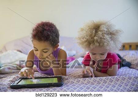 Stock Image - Mixed race sisters using digital tablets in bed. Fotosearch
