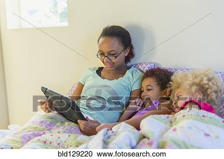 Stock Image - Mixed race sisters using digital tablet in bed. Fotosearch