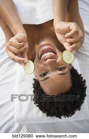 Stock Image - Portrait of smiling Black woman holding cucumber slices. Fotosearch