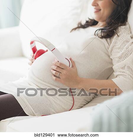 Stock Image - Pregnant Hispanic woman holding headphones on belly. Fotosearch