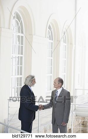 Stock Image - Senior Caucasian businessmen shaking hands on balcony. Fotosearch