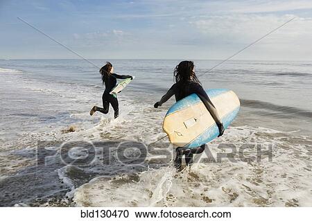 Stock Image - Surfers carrying boards in waves. Fotosearch