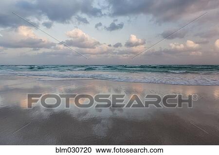 Stock Image - Clouds and sky reflected in still beach surf at sunrise. Fotosearch