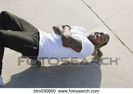 Stock Image - African American man laying on skateboard on city street. Fotosearch