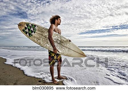 Stock Photography - Caucasian man holding surfboard in waves on beach. Fotosearch