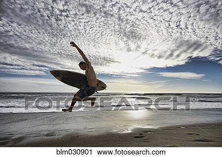 Stock Image - Caucasian man jumping with surfboard on beach. Fotosearch