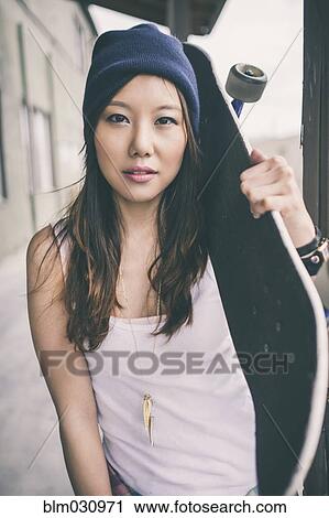 Stock Image - Korean woman carrying skateboard on city street. Fotosearch