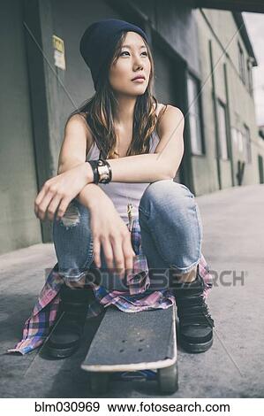 Stock Photo - Korean woman sitting on skateboard on city street. Fotosearch